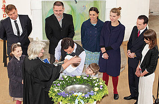 Eine Pfarrerin tauft ein Kind. Die Familie und die Taufpaten stehen mit am Taufbecken.